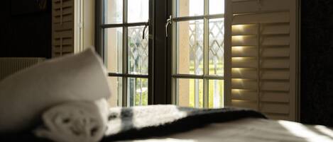 A bed with a rolled towel, a window with blinds, and a view of greenery outside.