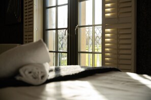 A bed with a rolled towel, a window with blinds, and a view of greenery outside. - 60 Lough Erne Golf Village · Escape Ordinary at Lough Erne! (Castle Hume/Lough Erne)