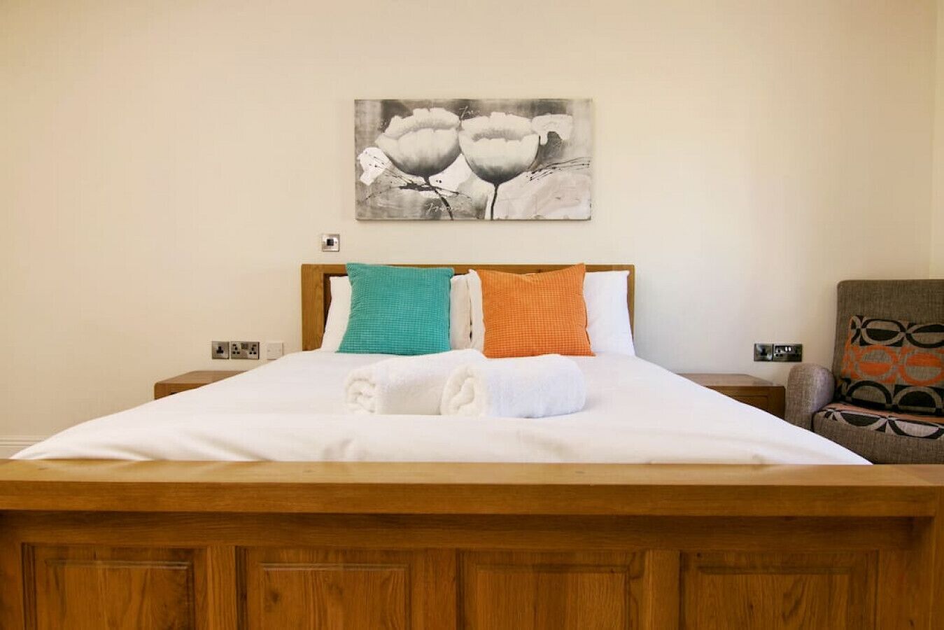 A neatly made bed with white linens and two pillows, a wooden headboard, and a painting of flowers on the wall.