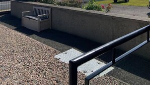 Een balkon met uitzicht op zee, een geparkeerde auto en een gazon met een bankje.