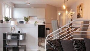 Een moderne woonkamer met een grijze bank, kussens met een zebraprint, een trap met witte leuningen en een keukenhoek met witte kastjes en een zwarte vaatwasser.