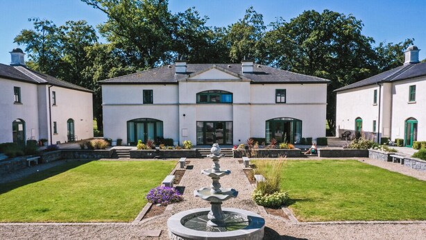 A large, white building with a central entrance, surrounded by a well-maintained garden and a fountain.