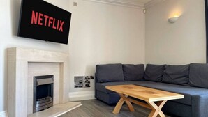 Un salon moderne avec une cheminée, un canapé gris, une table basse en bois et un téléviseur fixé au mur qui affiche « NETFLIX ».