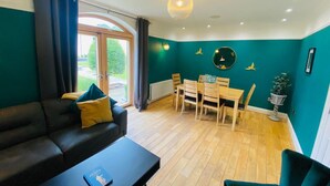 Una sala de estar moderna con una pared verde oscuro, piso de madera, un sofá negro, una mesa de comedor con sillas y una lámpara de techo.