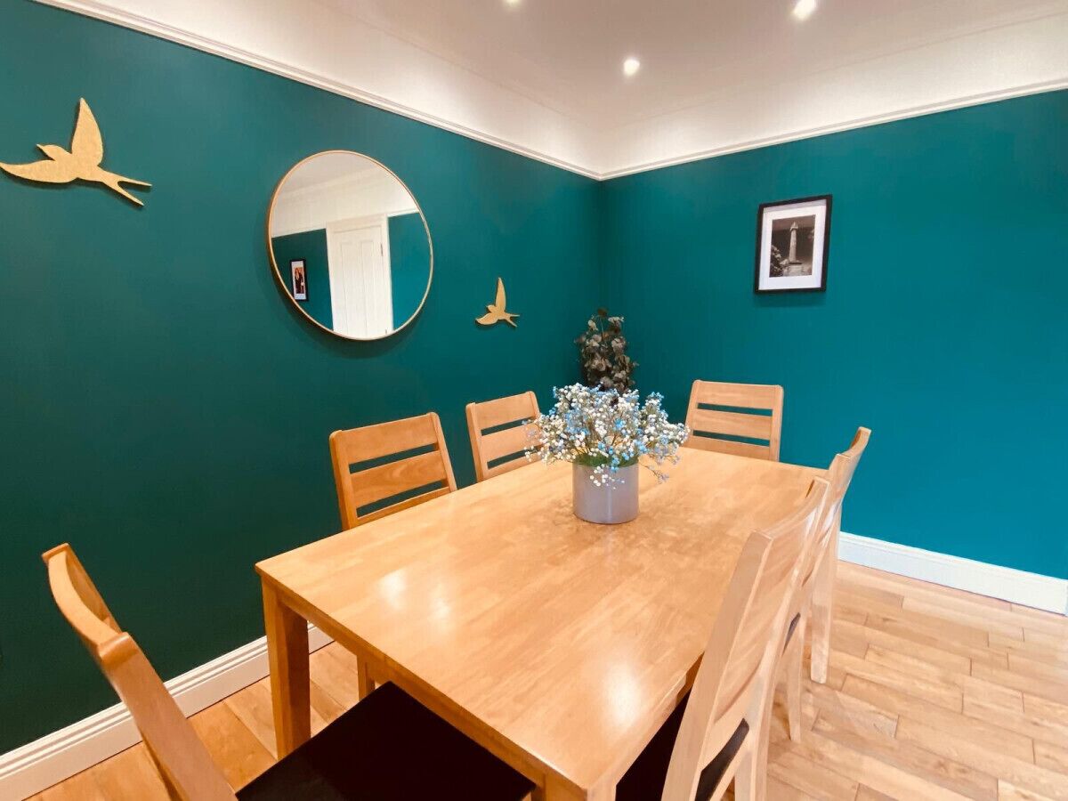Un coin repas avec une table et des chaises en bois, des murs de couleur turquoise, un miroir rond et un vase rempli de fleurs.