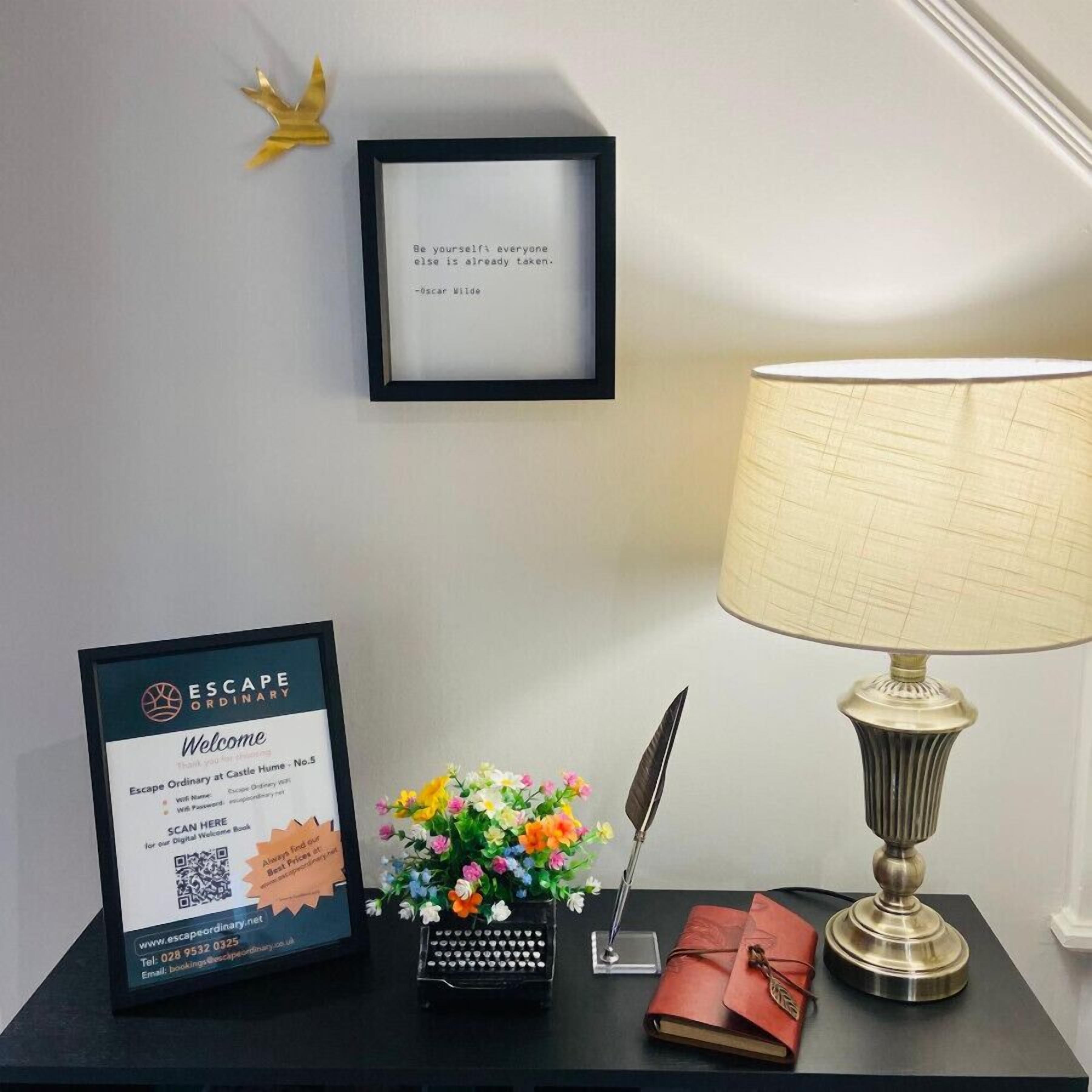 A framed welcome sign, a desk with a lamp, a typewriter, a notebook, and a vase of flowers.