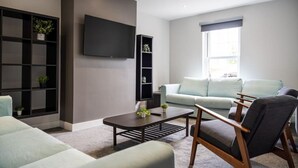 A modern living room with a sofa, armchairs, a coffee table, a TV mounted on the wall, and built-in shelving with plants.