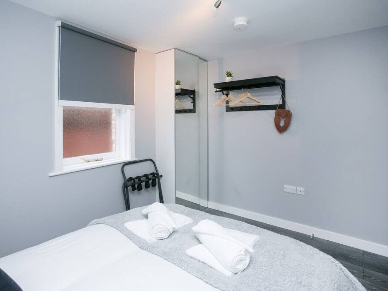 A modern bedroom with a bed, a window with blinds, a mirror, a towel rack, and a small shelf with a plant.