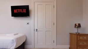Et værelse med en seng, en trækommode, en dør og et vægmonteret fjernsyn, der viser Netflix-logoet.