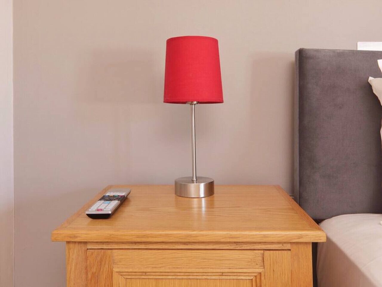 Une table de chevet en bois, avec une lampe rouge, une télécommande et une tête de lit grise.
