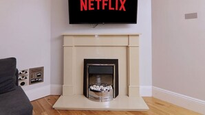 En stue med pejs, et tv monteret over den, der viser Netflix-logoet, og en grå sofa.