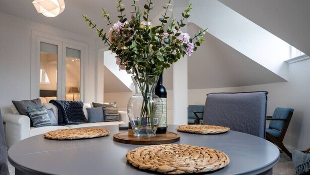 Un salon moderne avec une table ronde, des sets de table en fibres naturelles, un vase rempli de fleurs et une bouteille de vin.