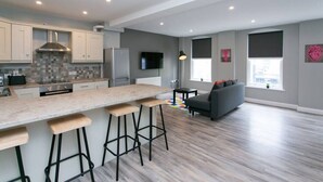 A modern kitchen with a central island, bar stools, and a grey sofa in a living area with a large window. - University Road 1 · Spacious & Modern + Central Location|Couples Break (Belfast)