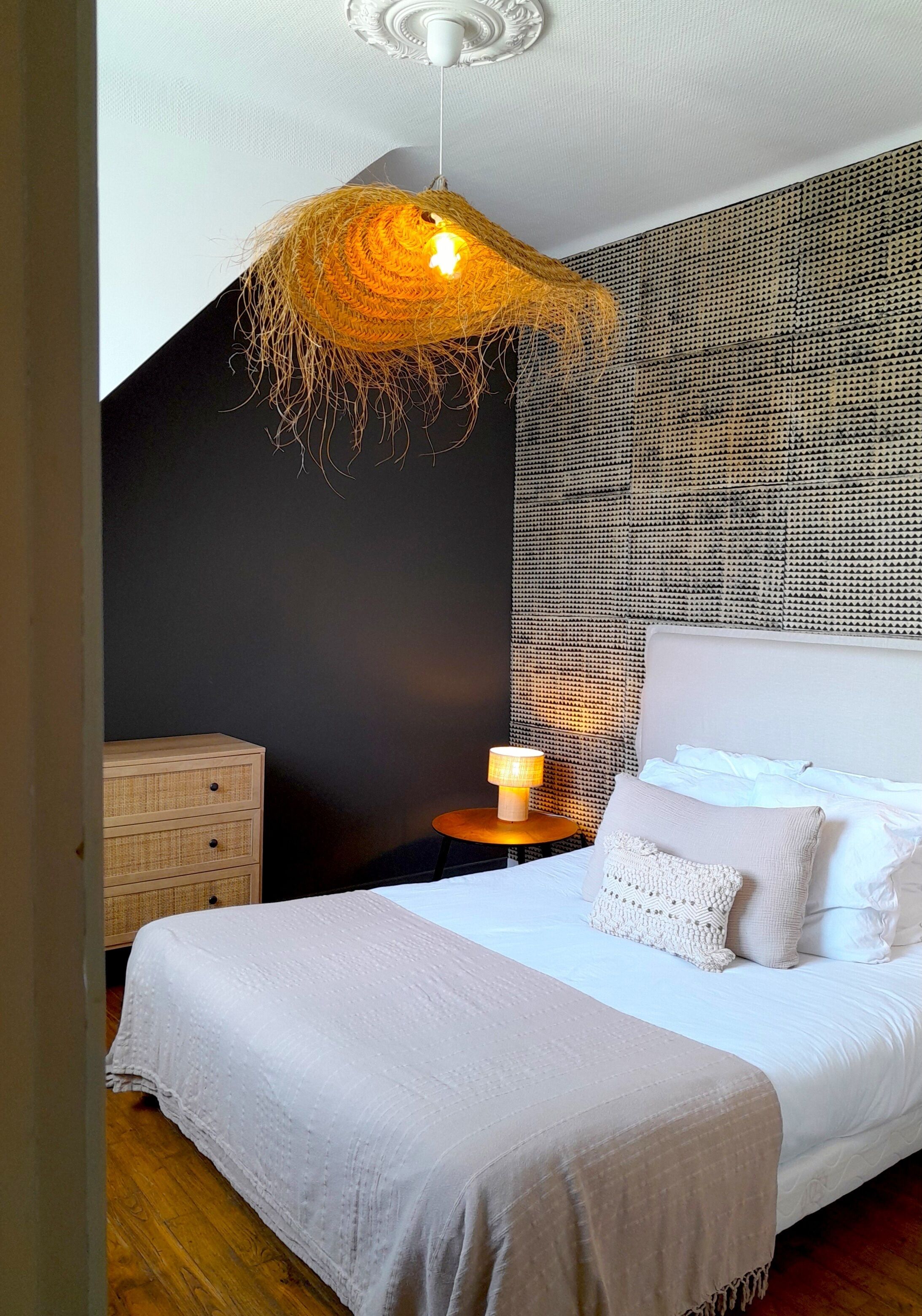 Ein Schlafzimmer mit einem großen Bett, einem Nachttisch mit Lampe, einer gewobenen Pendelleuchte und einer strukturierten Wand.
