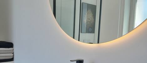 A modern bathroom with a black sink, a round mirror, and a small black trash bin.