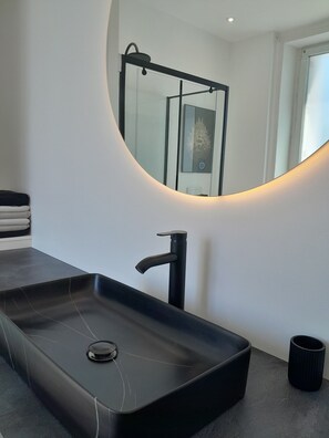 Ein modernes Badezimmer mit einem schwarzen Waschbecken, einem runden Spiegel und einer kleinen schwarzen Mülltonne.