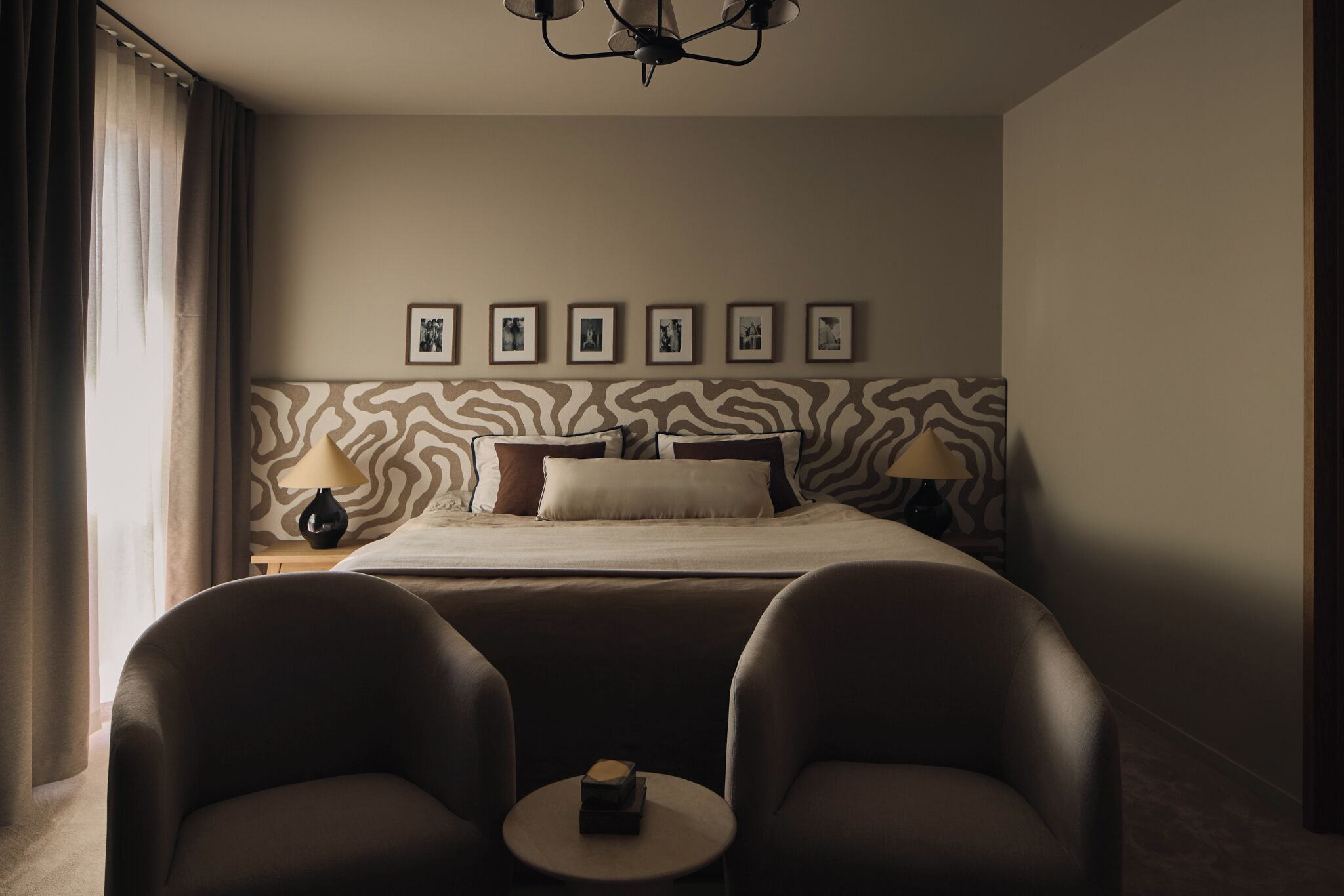 Habitación de hotel con cama, dos sillas, una mesita, lámpara y un muro con cuadros.