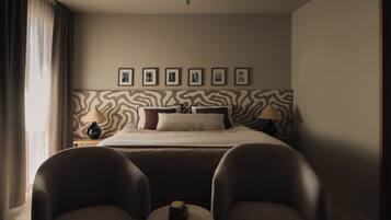 Habitación de hotel con cama, dos sillas, una mesita, lámpara y un muro con cuadros.