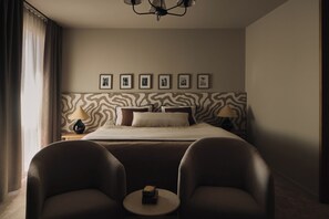 Habitación de hotel con cama, dos sillas, una mesita, lámpara y un muro con cuadros.