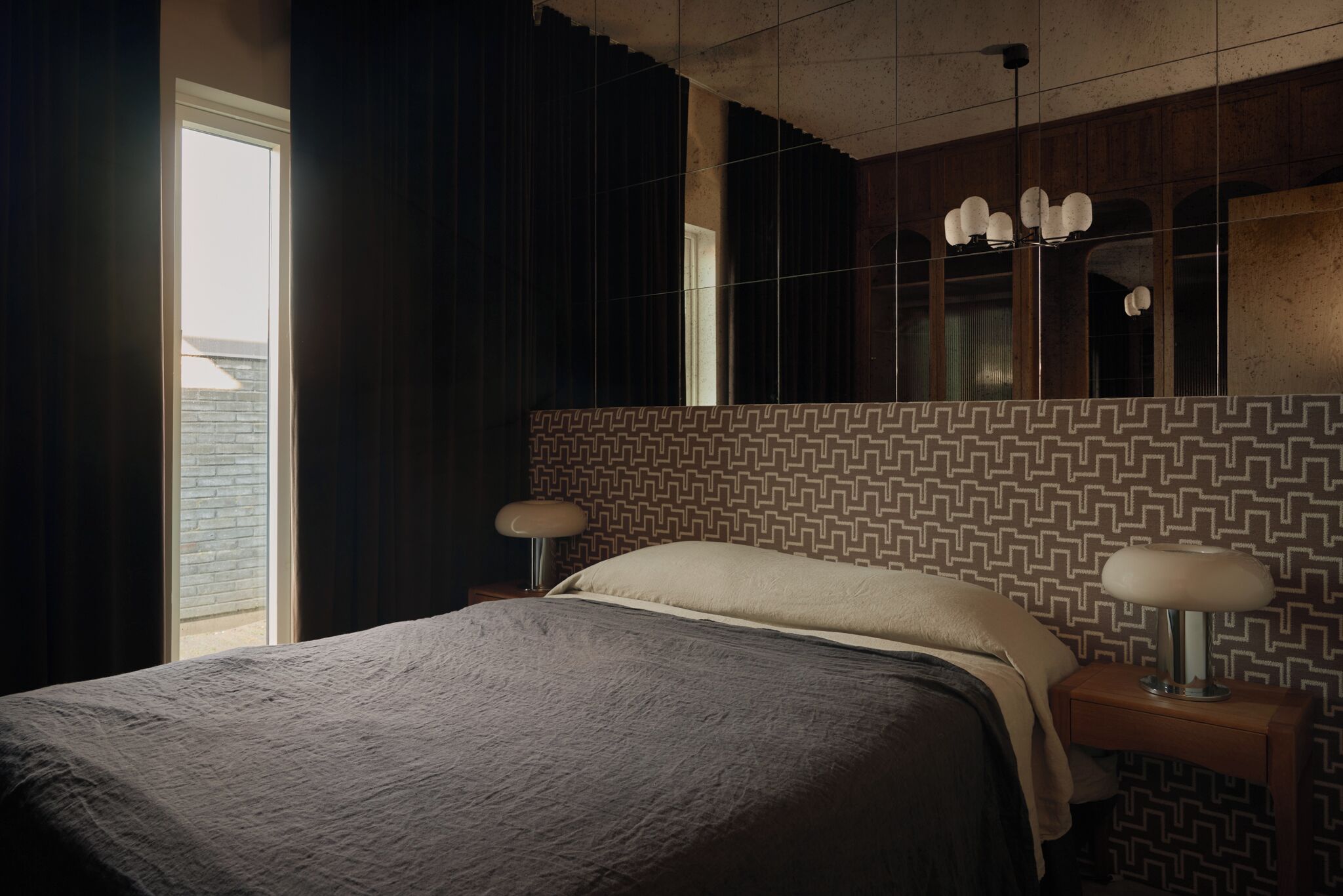 A hotel room with a bed, a nightstand, a lamp, a mirror, and a window with curtains.