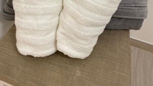 A pair of white slippers on a wooden surface, with a folded grey towel in the background. - Channel Suite - 2 (Sinalunga)