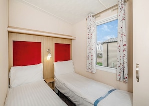 Quarto com duas camas, janela com cortinas florais, luminária de cabeceira e vista para o exterior.