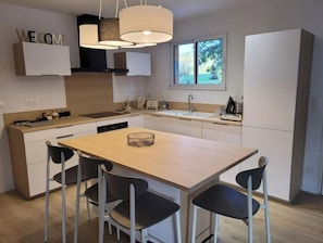 A modern kitchen with a central island, four chairs, a sink, and a window with a view of greenery.