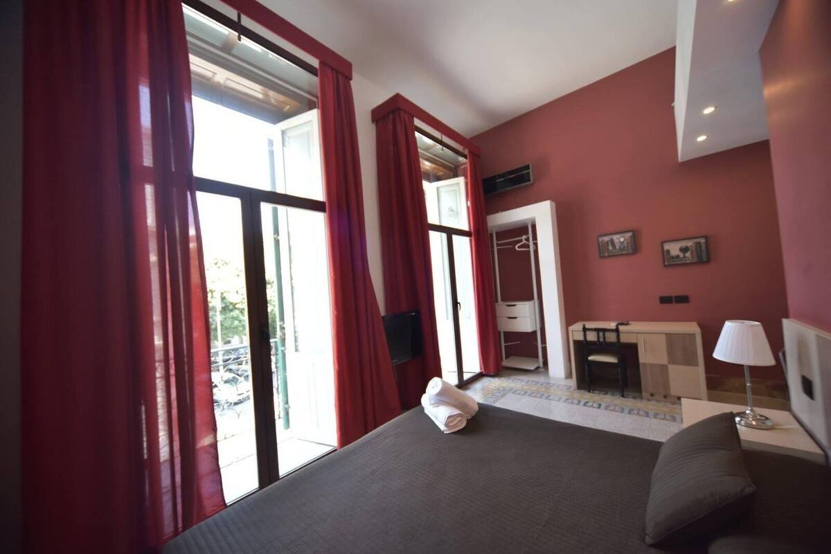 A hotel room with a bed, a desk, a chair, a wardrobe, and a window with red curtains.