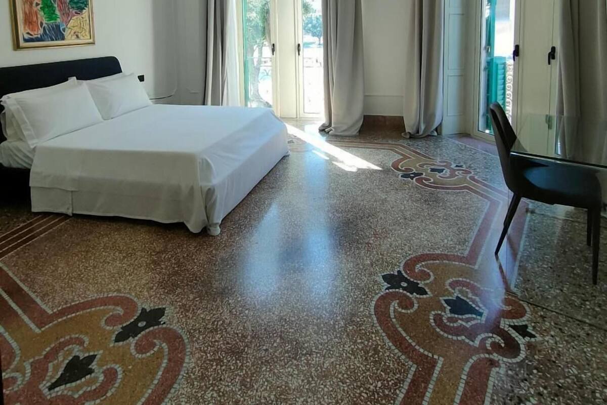 A hotel room with a bed, a chair, a large window with curtains, and a colorful rug.