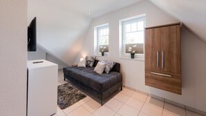 A modern loft bedroom with a loft bed, a grey sofa, a wooden wardrobe, and a TV mounted on the wall. - Vacation home "Juist" with 105sqm - in the vacation park Harkebrügge (Barßel)