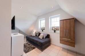 A modern loft bedroom with a loft bed, a grey sofa, a wooden wardrobe, and a TV mounted on the wall.