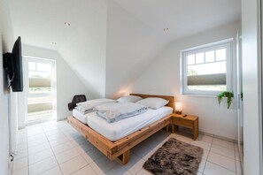 Quarto com cama de madeira, mesa de cabeceira, televisão na parede e janela com planta.