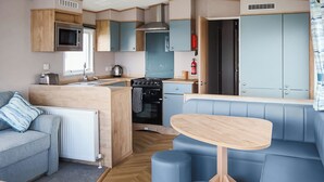 Eine kompakte Küchenzeile mit Mikrowelle, Toaster und Wasserkocher, ein Essbereich mit Tisch und blauem Sitzmöbel sowie ein Sofa mit blauem Kissen.