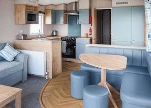 A compact kitchenette with a microwave, toaster, and kettle, a dining area with a table and blue seating, and a sofa with a blue pillow. - Otterton Premier Plus 3 (Budleigh Salterton)