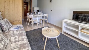 Una sala moderna con un sofá, una mesita redonda, un televisor sobre un mueble bajo y una zona de comedor con sillas y una mesa.
