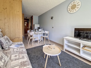 Uma sala de estar moderna com um sofá, uma mesa redonda pequena, uma TV sobre um suporte e uma área de jantar com cadeiras e uma mesa.