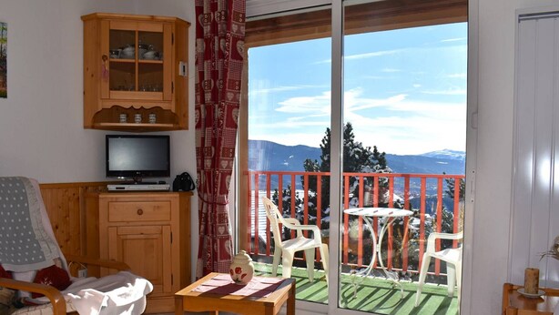 A cozy living room with a television, wooden furniture, and a balcony with a scenic mountain view. - Beautiful sunny 2-room apartment (Font-Romeu-Odeillo-Via)