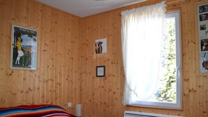 A room with wooden walls, a bed with a colorful striped blanket, a window with white curtains, and a poster on the wall. - Beautiful sunny 2-room apartment (Font-Romeu-Odeillo-Via)