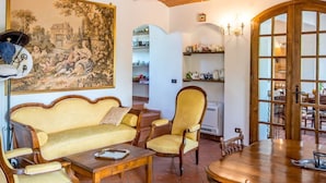 Una sala de estar acogedora con un sofá, sillones, una mesa de centro de madera y una mesa de comedor. Se observa un cuadro grande en la pared, estanterías con adornos y una puerta que da a otra habitación.