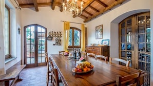 Un comedor con una mesa de madera dispuesta para cuatro personas, rodeada de sillas. Hay un frutero sobre la mesa y una araña colgando del techo.