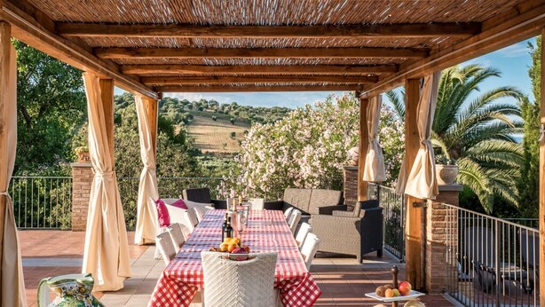 Un patio al aire libre con una mesa lista para comer, rodeado de sillas de mimbre y una pérgola.
