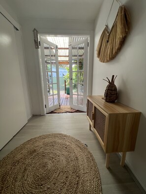 Een kamer met een groot geweven vloerkleed, een houten console en een deur naar een buitenruimte met een houten terras en begroeiing.