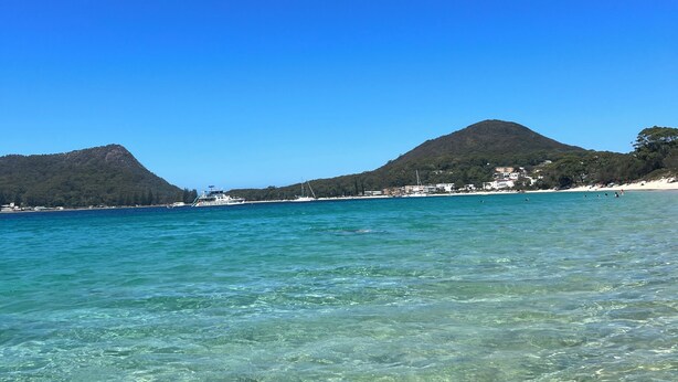 Un paysage côtier avec une eau turquoise et limpide, une plage de sable et une chaîne de montagnes en arrière-plan.