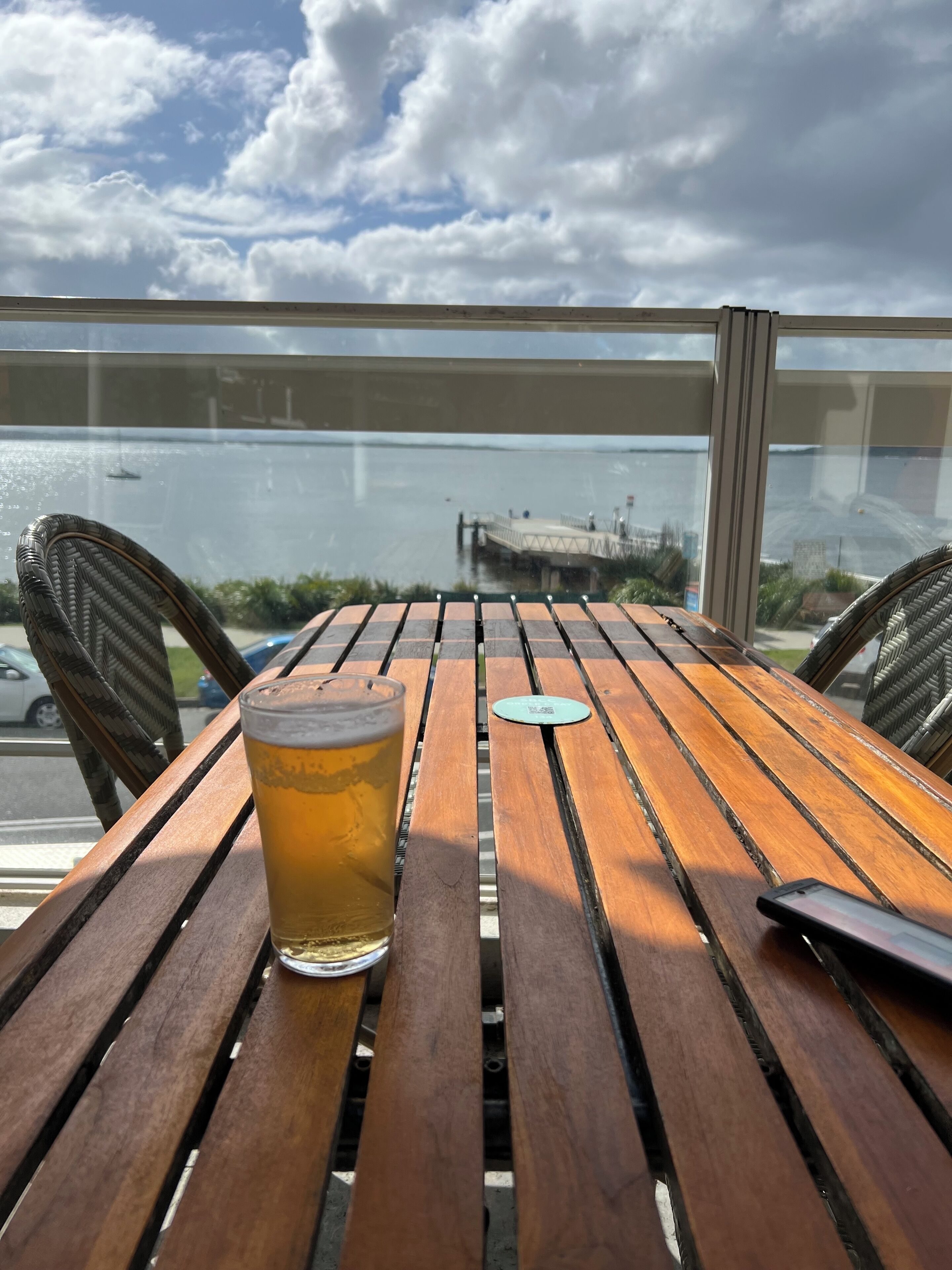 A wooden table with a glass of beer and a coaster, overlooking a body of water with a dock and a bridge in the distance.