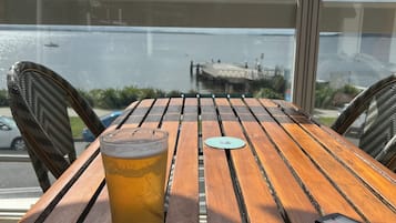 A wooden table with a glass of beer and a coaster, overlooking a body of water with a dock and a bridge in the distance.