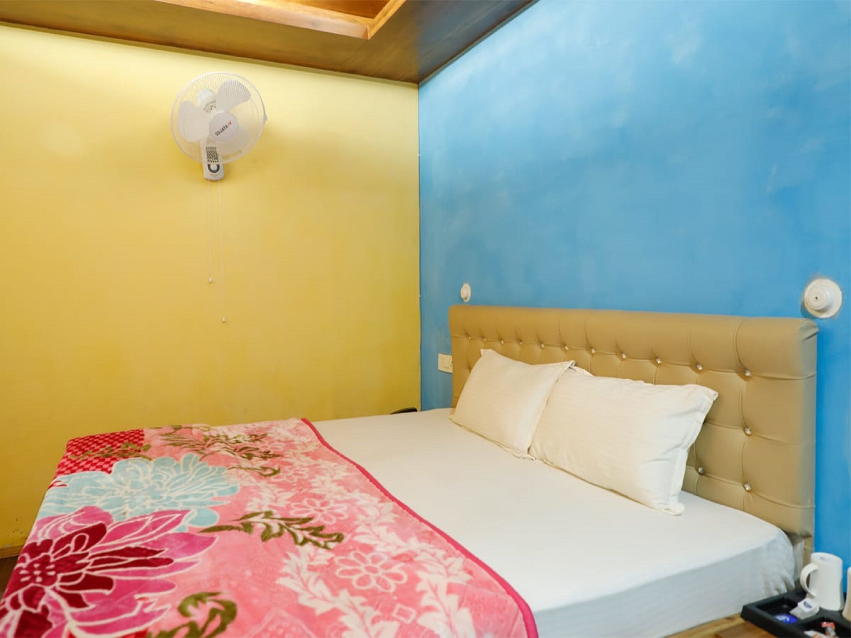 A bedroom with a bed, a bedside table with a cup, and a wall-mounted fan.