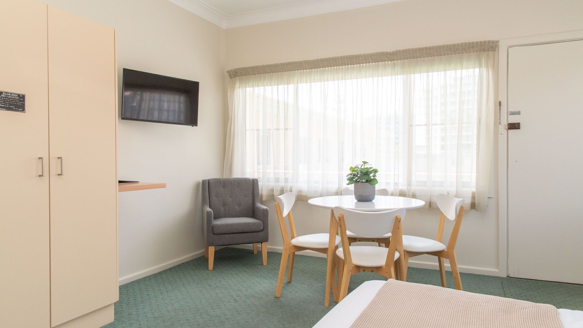 Quarto de hotel com cama, escrivaninha, cadeira, televisão fixada na parede e janela com cortinas finas.
