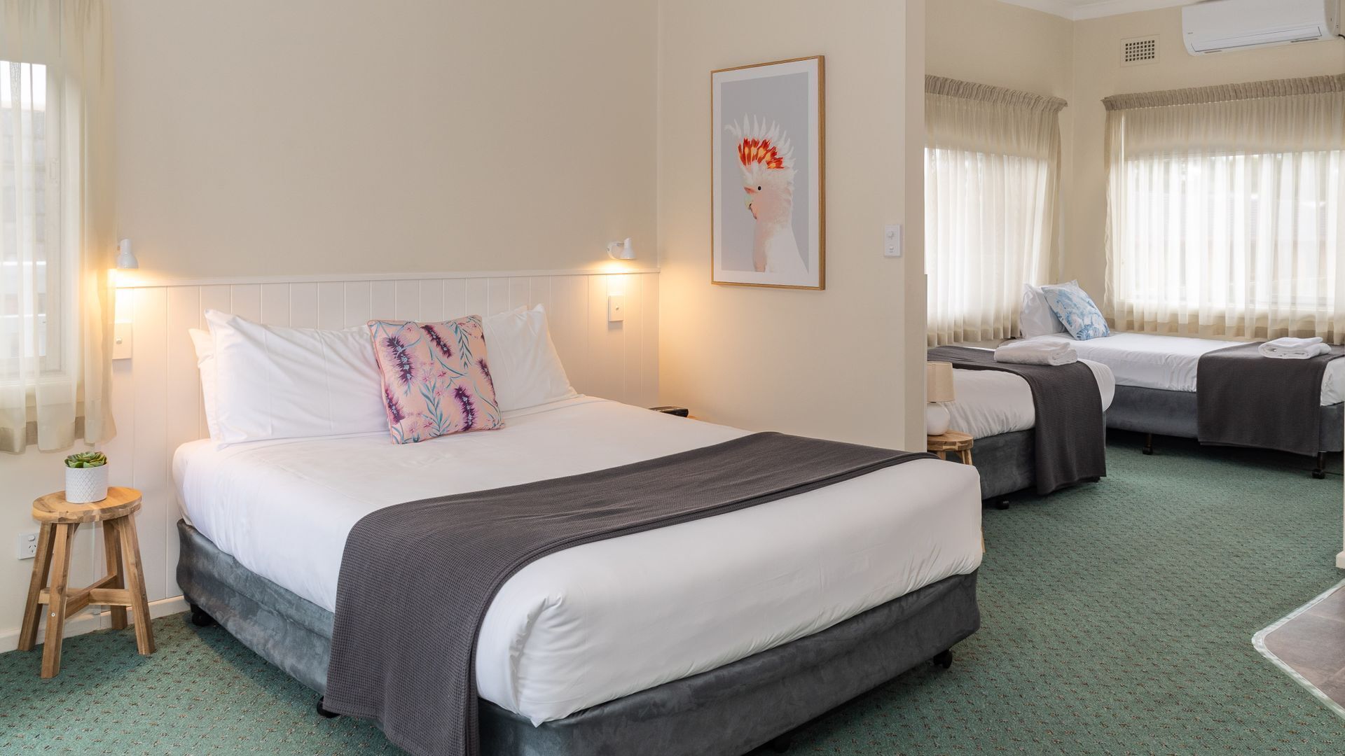 Een hotelkamer met twee bedden, een klein krukje, een ingelijst schilderij en een groen tapijt.