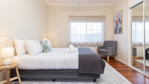 A bedroom with a bed, a chair, a bedside table, a lamp, and a window with sheer curtains. - Jasmine Lodge in the heart of Forster (Forster)