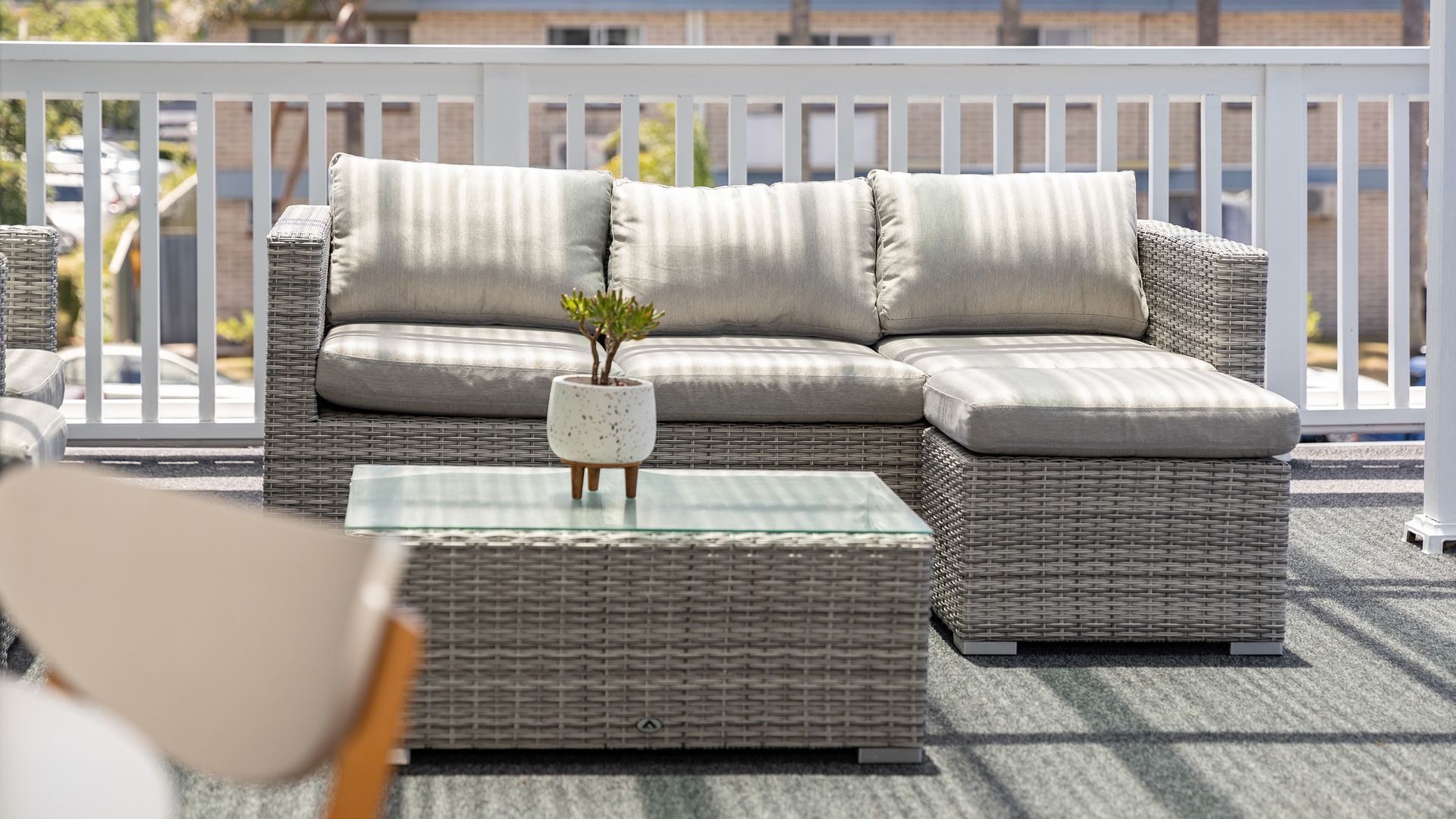 Een rieten bank met kussens en een glazen salontafel met een plant, op een balkon met een witte balustrade.